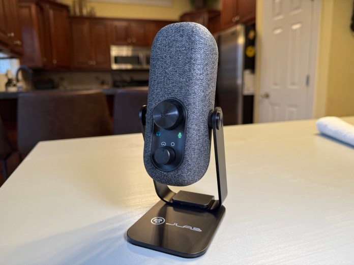 Close-up of the JLab Go Talk USB microphone on a sturdy stand, featuring a sleek design with knobs and buttons for volume and gain control. The microphone is set against a kitchen background.