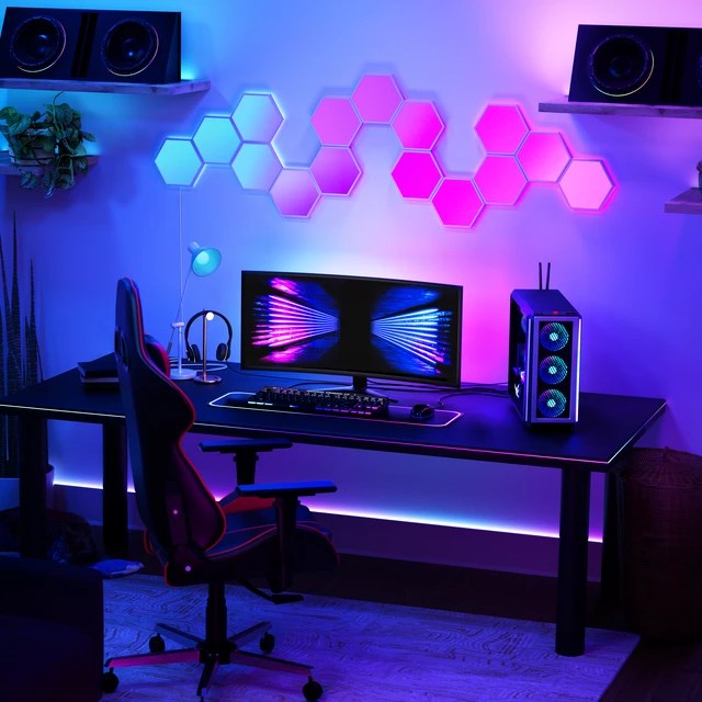 A modern gaming setup featuring a sleek desktop monitor, gaming keyboard, and a colorful illuminated background with hexagon panels. The setup includes a gaming chair and ambient lighting, creating a dynamic atmosphere suitable for gaming.