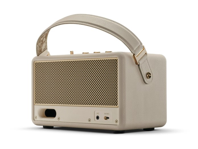Cream colored portable Bluetooth speaker with a textured surface, featuring a gold mesh grille, vintage-style knobs, and a carry strap.