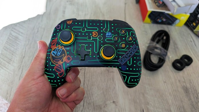 Close-up of a PowerA Pac-Man Neon Arcade Controller, showcasing its RGB LED lighting and retro design, held in a hand over a wooden surface.