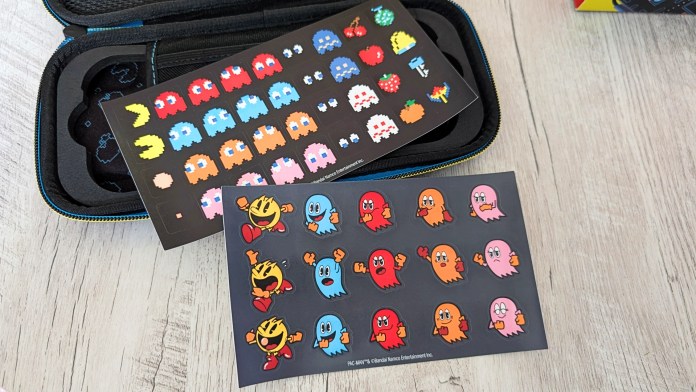 A close-up view of two sticker sheets featuring colorful Pac-Man characters, including ghosts, fruits, and Pac-Man himself, displayed on a textured surface next to a gaming case.