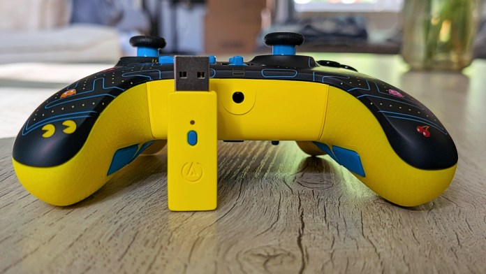 A PowerA Pac-Man wireless controller for Xbox, featuring a vibrant yellow design with classic Pac-Man artwork, positioned next to a small USB adapter on a wooden surface.