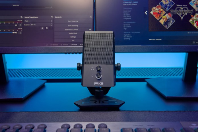 A RIG gaming microphone on a desk with dual monitors in the background, featuring a vibrant blue ambient light.