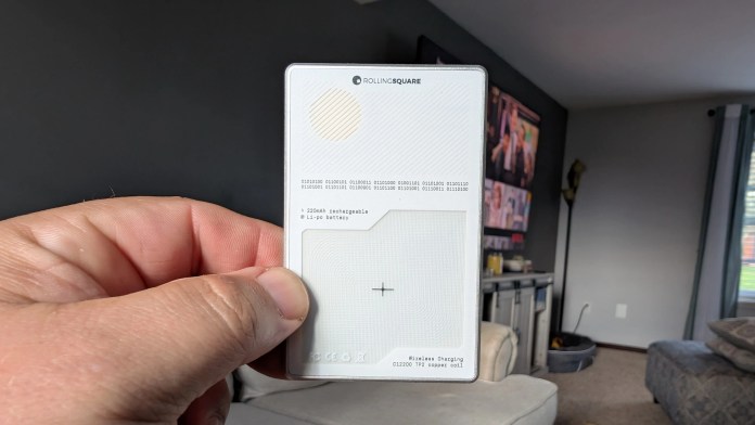 A person holding a Rolling Square AirCard Pro Bluetooth tracker in their hand, with a living room background featuring a TV and furniture.