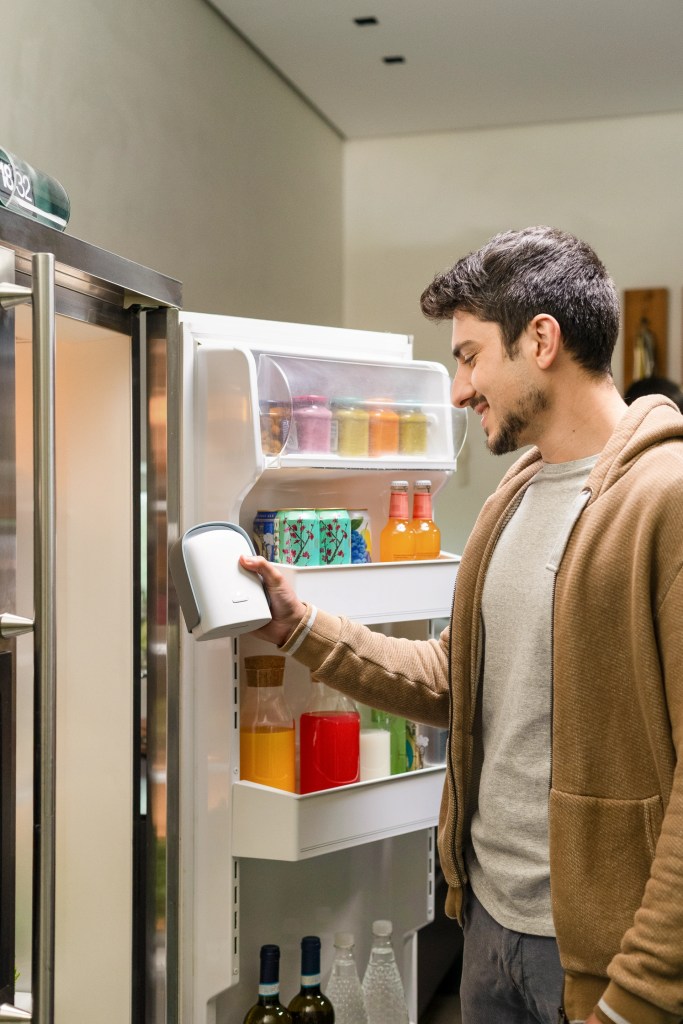 A man standing in front of an open refrigerator, holding the Shelfy Lite air purifier with various colorful bottles and jars visible inside.