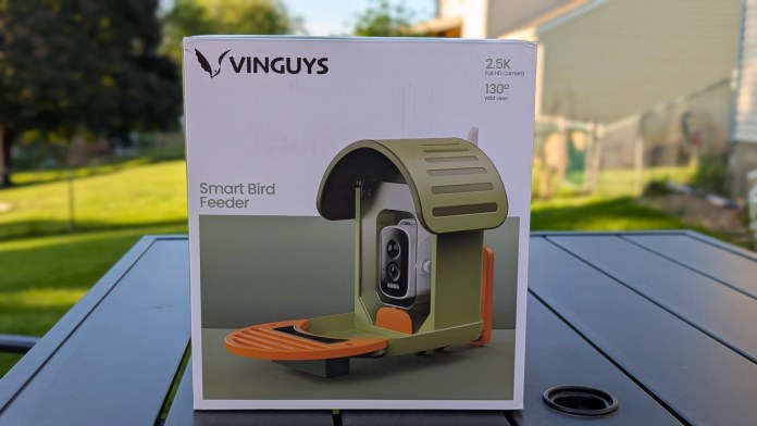 Box packaging for the Vinguys Smart Bird Feeder V-Feeder featuring a 2.5K Full HD camera and a 130° wide view with a green and orange design, set on a table outdoors.