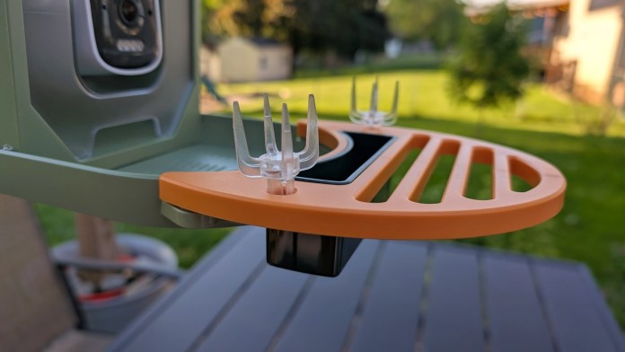 Close-up of the feeding area of the Vinguys Smart Bird Feeder V-Feeder, showcasing its orange platform with clear feeding spikes and integrated camera, set against a blurred outdoor background.