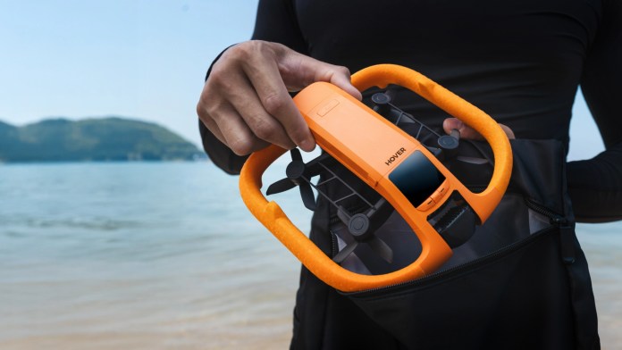 A person holding the HOVERAir AQUA, a waterproof self-flying camera designed for water activities, against a serene beach backdrop.