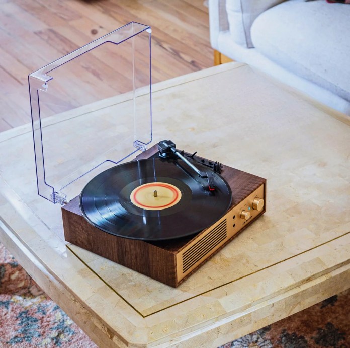 A Crosley record player showcasing a vinyl on a table, with a transparent dust cover partially closed, surrounded by a cozy interior setting.