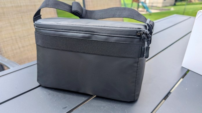 A black, square-shaped carrying case designed for portable electronics, featuring a sturdy zipper and a handle, placed on a wooden table.