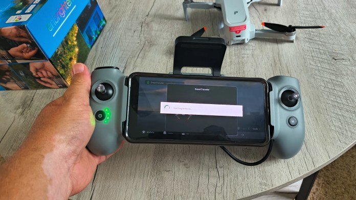 A close-up view of a hand holding the AT2-RC remote controller for the Potensic ATOM 2 drone, featuring a smartphone mounted on the controller with a user interface displayed. The background shows the drone and its packaging on a wooden surface.