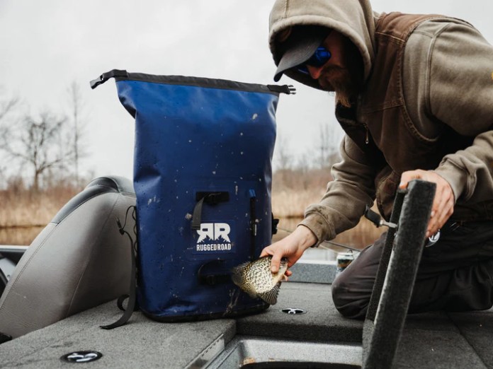 A person wearing a hat and sunglasses is handling a fish while sitting next to a blue Rugged Road cooler on a boat.
