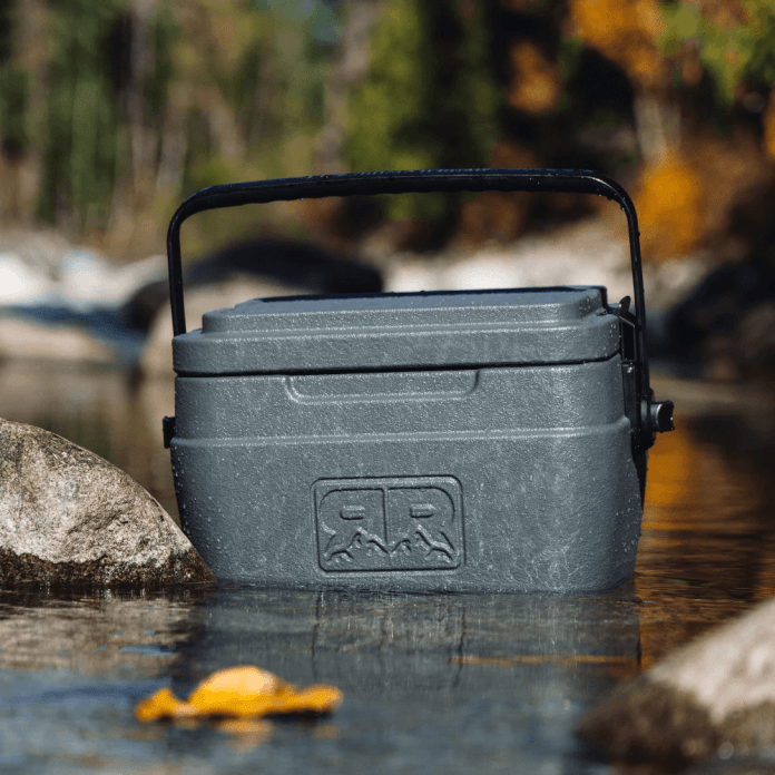 A Rugged Road cooler partially submerged in shallow water, surrounded by rocks and trees, showcasing its durability and floatability.