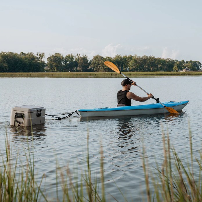 A person kayaking on a calm body of water, towing a lightweight cooler behind them.