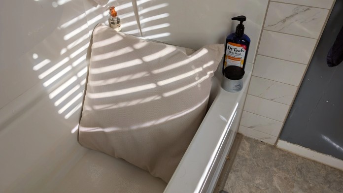 A soft, neutral-colored bath pillow resting in a bathtub, surrounded by soft light from shadows on the wall. A bottle of Dr. Teal's bath wash is placed next to the pillow on the tub's edge.