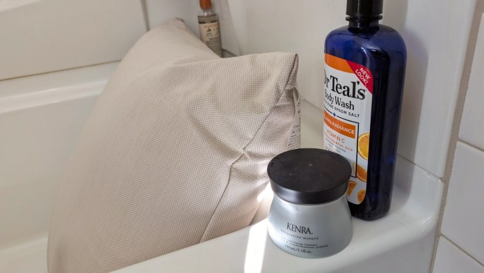 A beige bath pillow resting against a tub, accompanied by bottles of Dr. Teal's body wash and Kenra nourishing masque on the ledge.