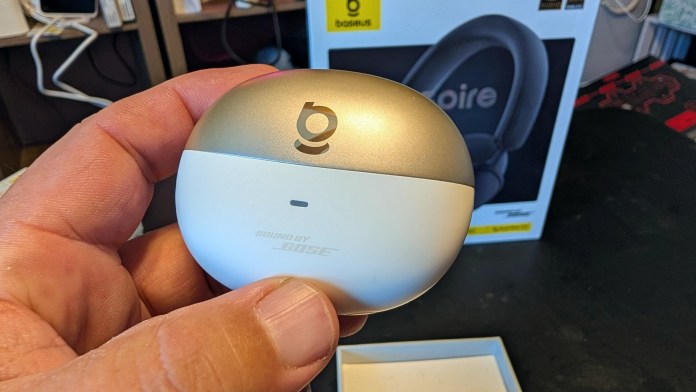 A close-up of a person's hand holding a white and gold charging case for headphones, with the logo visible on the top. In the background is a display box for Baseus Inspire headphones.