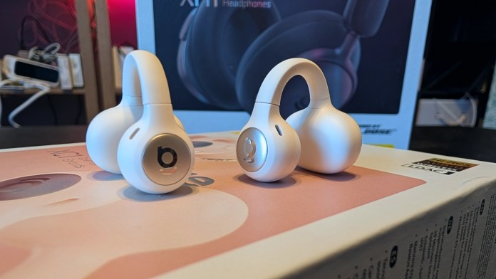 Close-up of two Baseus Inspire XC1 Open-Ear Earbuds in white, displayed on a product box, with the Baseus Inspire XH1 headphones visible in the background.