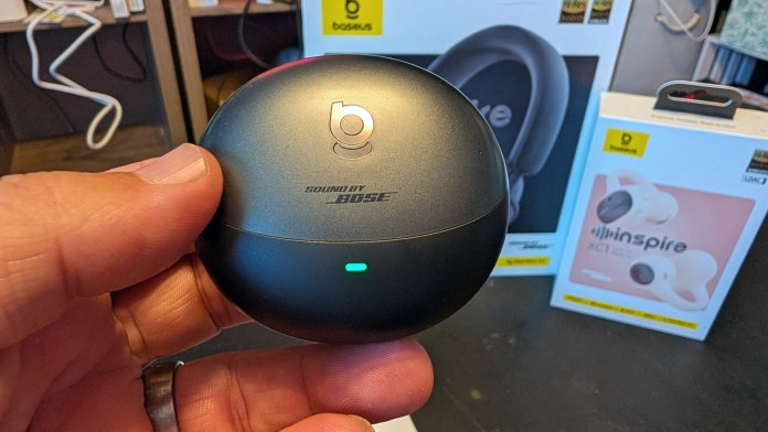 A person holding a charging case for wireless earbuds branded 'Sound by Bose', with blurred images of other audio products in the background.