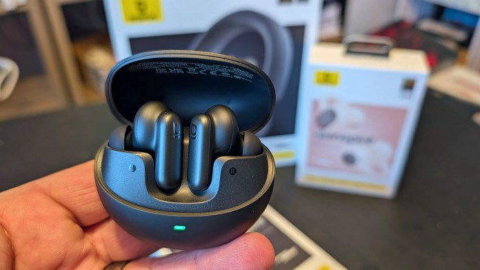 A hand holding a black charging case with two wireless earbuds inside, surrounded by product packaging in the background.