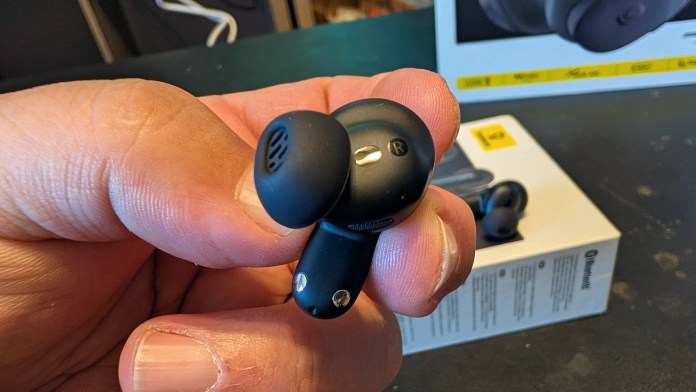 A person holding a black wireless earbud, showcasing its design and details, with a product box visible in the background.