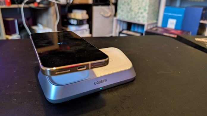 A UGREEN MagFlow 3-in-1 Magnetic Wireless Charger with a smartphone on top, displaying its charging status of 87%. The charger features a sleek design and is positioned on a dark surface with various items in the background.