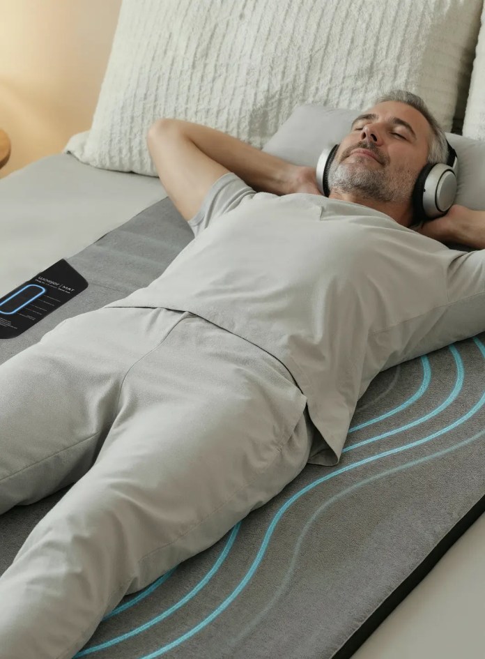 A man relaxing on a Woojer MAT mattress topper, wearing headphones and enjoying a calm moment, with soft bedding and a peaceful bedroom ambiance.