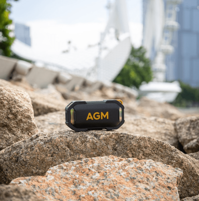 A portable AGM speaker resting on a rocky surface with a blurred outdoor background featuring modern architecture.
