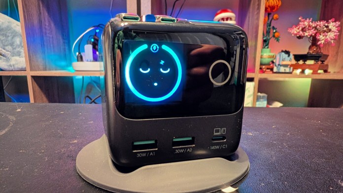 A compact black charging station featuring multiple USB ports and a digital display showing a smiling face icon, set on a flat surface with a blurred background of shelves and decorations.