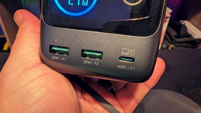 A close-up view of a hand holding a device with multiple USB ports labeled with power specifications: two 30W ports (A1, A2) and one 140W port (C1), alongside a digital display showing power output.