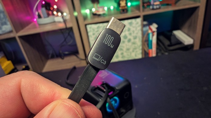 A close-up view of a hand holding a USB-C cable labeled '100W' against a blurred background with various objects on shelves.