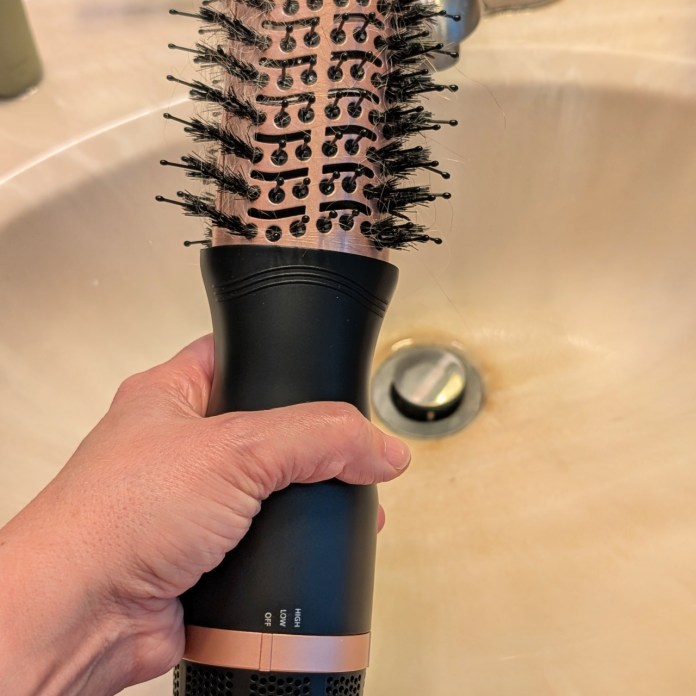 A hand holding a round brush with bristles over a bathroom sink.