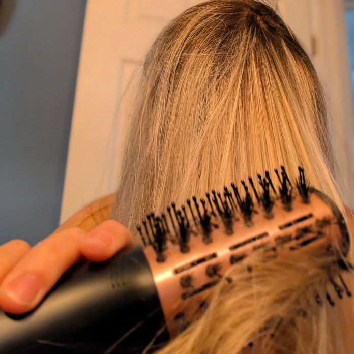 A person using a round brush to style long, blonde hair in a bathroom setting.