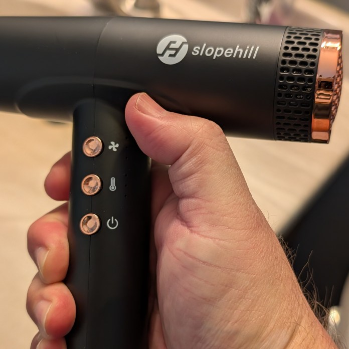 A person holding a black hair dryer with a logo and three control buttons, set against a blurred background.