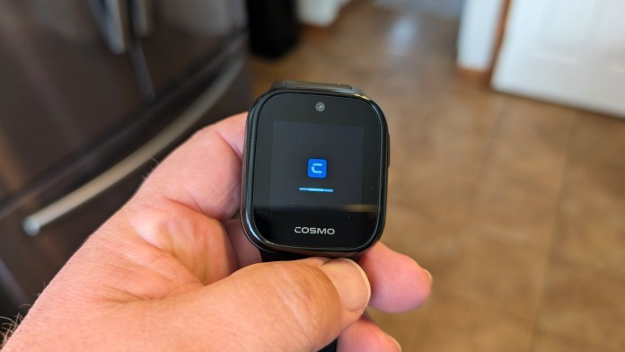 A hand holding a COSMO smartwatch displaying a blue loading screen in a kitchen setting.