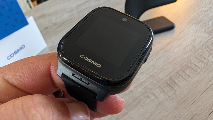 A hand holding a black smartwatch with a square face displaying the brand name 'COSMO', set on a wooden surface.