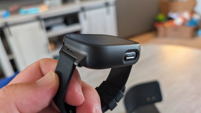 A close-up of a smartwatch being held, showcasing its side view with a USB charging port and strap.