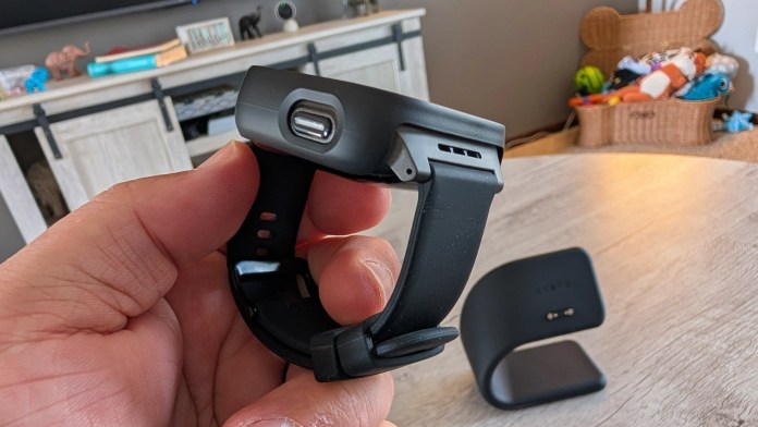 A person holding a black smartwatch with a USB charging port, displaying the underside of the device, and a charging stand in the background.