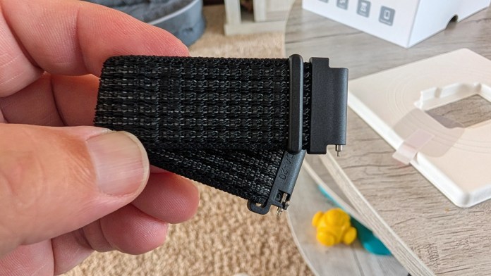 A close-up of a person holding a black woven strap with a plastic attachment, set against a blurred background of a table and other objects.