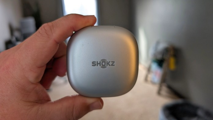 A person holding a silver charging case for Shokz wireless headphones, with a blurred background of a room.