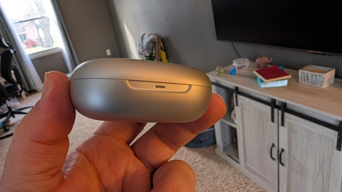 A person holding a silver wireless earbuds charging case in a living room setting.