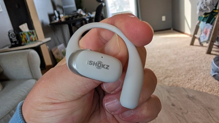 A person holding a pair of gray Ishokz wireless headphones with a background featuring a home office setting.