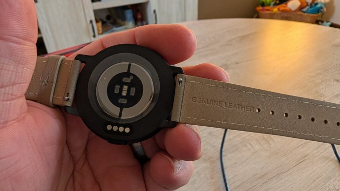 A person is holding a smartwatch with a black casing and a genuine leather strap. The back of the watch features sensors and a circular design.