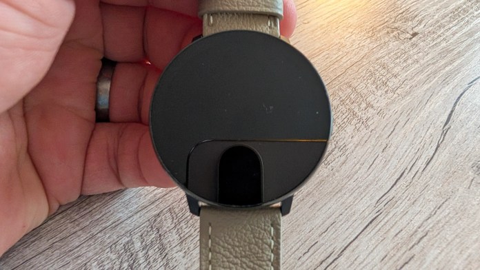 A person holding a circular smartwatch with a black screen and a leather strap, placed on a wooden surface.