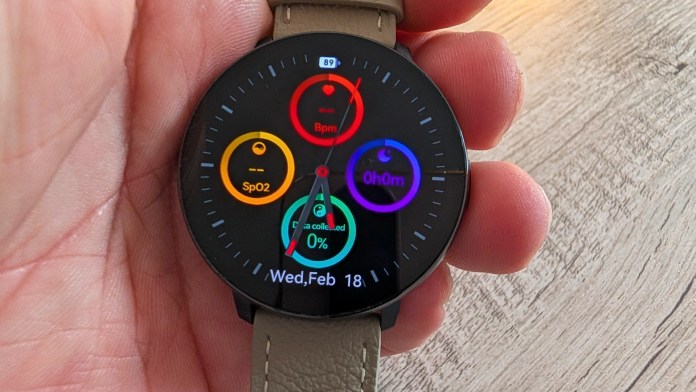 A close-up of a smartwatch displaying health metrics including heart rate (Bpm), blood oxygen level (SpO2), and data collected percentage. The watch is held in a hand against a wooden surface.