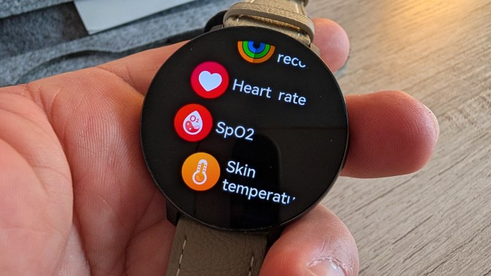 A close-up of a hand holding a smartwatch displaying health metrics including heart rate, SpO2, and skin temperature.