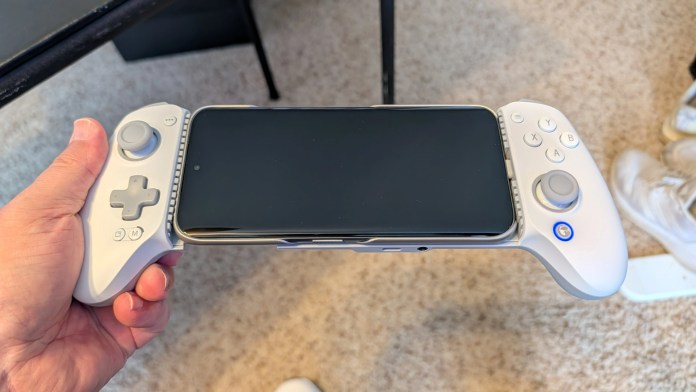 A person holding a white gaming controller with a smartphone attached to it, featuring a blank screen, on a carpeted surface.