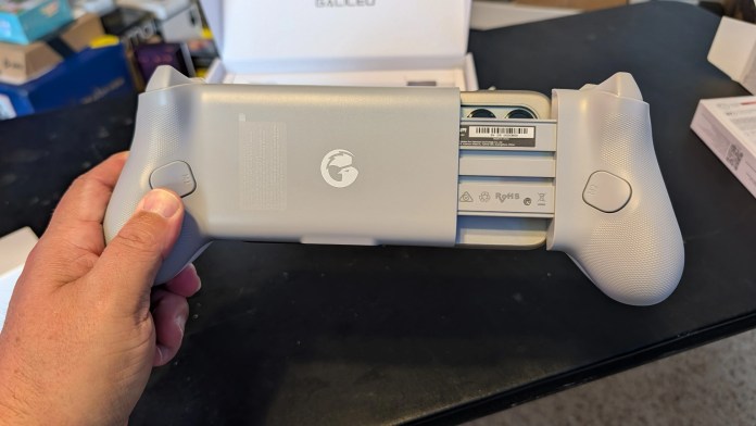 A person holding a gray gaming controller with visible buttons and a logo, positioned on a black table with a partially open box in the background.