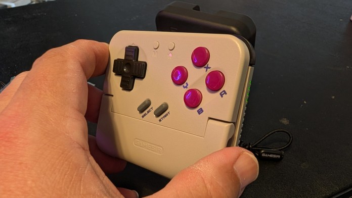A hand holding a retro-style game controller featuring a directional pad, colorful action buttons, and labeled start/select buttons, set on a dark background.