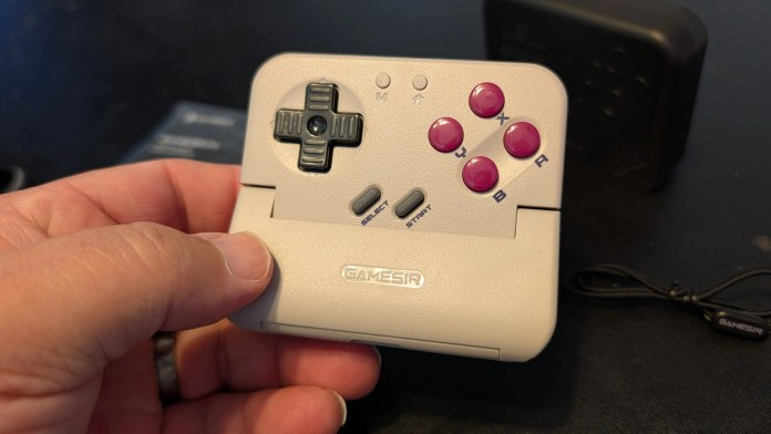 A person holding a retro-style gaming controller with a directional pad and buttons labeled A, B, X, and Y. The controller is gray with pink accents and features a start and select button.
